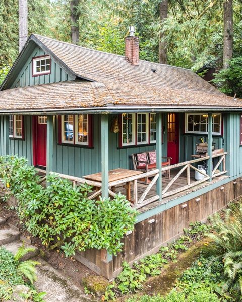 Green wooden cabin with a wraparound porch surrounded by forest ferns